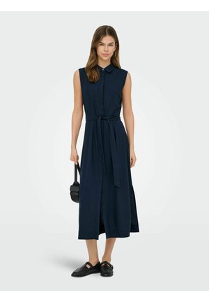 Nő ujjatlan tengerészkék midi ingruhával, derékpánttal, kezében kis fekete kézitáskával, és fekete balerina cipővel a lábán.