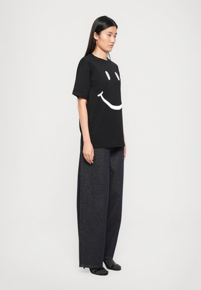 Svart bomull T-skjorte med et stort hvitt smilende ansikt grafikk. Kombinert med mørke, løstsittende denim-bukser og svarte sko.
