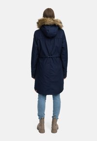 Mornarska parka z umetnim krznom na kapuci, zategovanjem v pasu, dolgimi rokavi in stranskimi žepi. Obuta z svetlo modrimi kavbojskimi hlačami in bež gležnjarskimi čevlji.