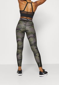 Nainen, joka on pukeutunut mustaan urheilu-liivimekkoon, camo-kuosiset legginsit, joissa on "PRO" vyötärö, ja mustiin Nike-tennarit, seisoo kädet ylhäällä.