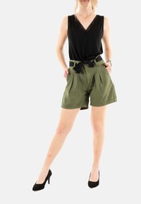 Top negro sin mangas con encaje, combinado con pantalones cortos de tiro alto verdes que presentan un cinturón y sutiles pliegues; terminado con tacones negros.