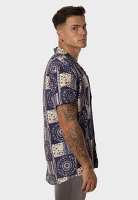 Camisa de manga corta en azul marino con patrones de paisley en crema y geométricos. Presenta un corte relajado, cuello abotonado y material de algodón suave.