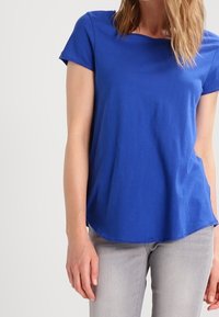 Blå kortärmad bomullstop med rundad halsringning och böjd nederkant. Slät textur; avslappnad passform, kombinerad med ljusgrå jeans.