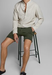 Svetle béžová halenka s třemi knoflíky a tmavě zelené šortky. Model sedí na barové stoličce, na nohou má černé sandály na klínku.