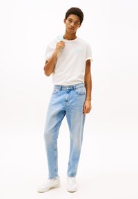 Hvid bomulds t-shirt med mintgrøn tekst, parret med lyseblå denimjeans. Modellen står med en lys jakke hængende over den ene skulder.