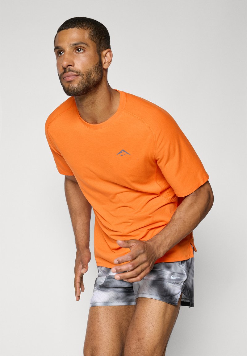 Camiseta atlética de manga corta de color naranja brillante con escote redondo, que presenta un pequeño logotipo azul. Combinada con pantalones cortos grises con patrón.