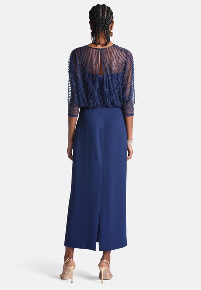 Femme aux cheveux tressés portant une robe sans manches bleu marine avec un overlay transparent orné de perles et des sandales à talons hauts dorées, debout de dos.