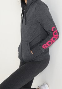 Grijze zip-up hoodie met roze logo op de mouw, voorzien van een kangoeroezak en een capuchon met trekkoord, gedragen met bijpassende donkere leggings.