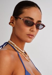 Maroon cat-eye sunglasses with dark lenses, intricate brown beaded necklaces, and a blue fabric top. Smooth skin with a natural finish.