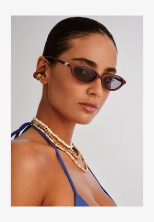 Maroonske sunčane naočale u obliku mačjih očiju s tamnim lećama, složene smeđe perle na ogrlicama i plava tkanina na gornjem dijelu. Glatka koža s prirodnim završetkom.