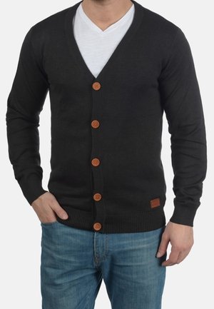 Homme portant un cardigan noir boutonné sur un t-shirt blanc à col en V et un jean bleu, une main dans la poche, sur un fond uni.