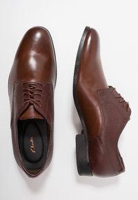 Chaussures habillées en cuir marron avec surfaces lisses et texturées, bout arrondi, design à lacets, et talon bas en caoutchouc noir.