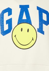 Bílá mikina s kapucí, na které je nápis "GAP" ve velkých modrých písmenech nad velkým žlutým smajlíkem. Materiál vypadá hladce a jemně.