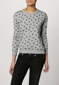 Felpa grigia con piccoli motivi neri, collo rotondo, polsini e orlo a coste, texture morbida, indossata con jeans scuri.
