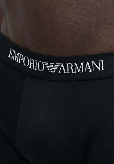 Gros plan d'une personne portant des sous-vêtements noirs Emporio Armani avec le logo et le nom de la marque visibles sur la ceinture.