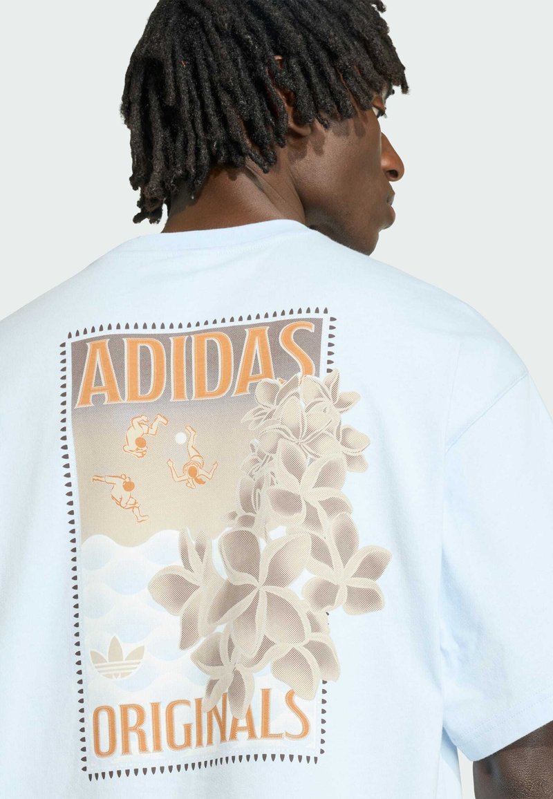 Person med korte dreadlocks iført en hvid Adidas Originals T-shirt med blomster og små dykkende figurer på ryggen.