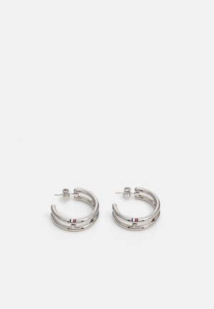 Boucles d'oreilles argentées à double cercle avec petites pierres claires et détail rayé rouge, blanc et bleu sur fond blanc.