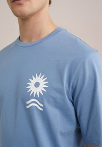 Világoskék pamut póló, elöl fehér nap és hullám grafikai tervezéssel. Kerek nyakú és rövid ujjú. Sima textúra.