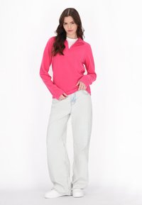 Donna che indossa una maglietta a maniche lunghe rosa brillante, jeans larghi blu chiaro e sneakers bianche, in piedi contro uno sfondo bianco.