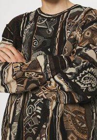 Sweter dziany łączący kolory brązowy, kremowy i czarny, z misternymi geometrycznymi wzorami i teksturowanymi detalami na całej tkaninie.