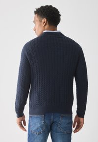 Pull en maille bleu marine avec un motif en câble texturé, doté d'un col rond classique et de poignets côtelés, associé à un jean en denim.