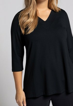 Femme portant un haut tunique noir avec un col en V, manches trois-quarts et cheveux blonds tombant sur son épaule, sur un fond uni.