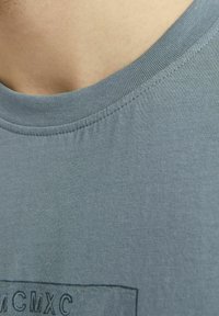 Tricou din bumbac gri, cu guler rotund, textură netedă și un design imprimat rectangular cu 'MCMXC' în centrul său.