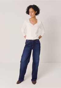 Weißes, geripptes Langarmoberteil mit V-Ausschnitt und dunkelblauen Schlagjeans. Das Model steht vor einem neutralen Hintergrund und trägt braune Knöchel-Stiefel.