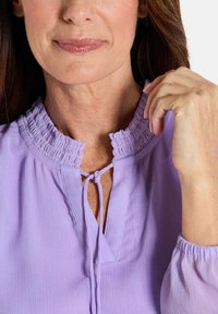 Vrouw in een lichtpaarse blouse met een gerimpelde kraag en strik, die met haar rechterhand de kraag aanraakt.