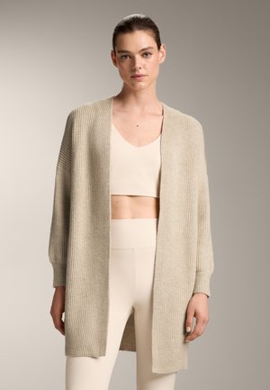 Cardigan beige a coste con apertura frontale, vestibilità ampia e maniche lunghe. Indossato sopra un top corto abbinato e leggings a vita alta.