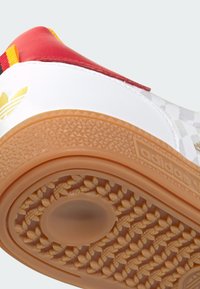 Gros plan de la semelle d'une chaussure Adidas avec une bande de roulement en caoutchouc marron clair, dessus blanc, talon rouge et logo Adidas doré sur l'étiquette du talon.
