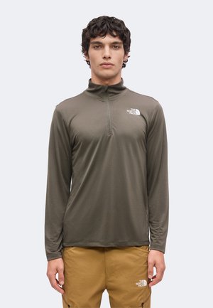 Młody mężczyzna w oliwkowej bluzie z długim rękawem i półzamkiem oraz beżowych spodniach, obydwa z logo The North Face, stoi na jasnym tle.