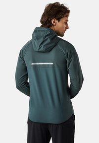 Giacca a cappuccio da uomo in blu scuro, realizzata in tessuto morbido e elastico. Presenta un pannello con logo riflettente sulla parte superiore della schiena e maniche aderenti.