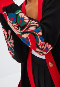 Cárdigan negro con acentos rojos, con patrones florales en azul, verde y blanco. Textura suave y cierre de botones en la parte frontal.
