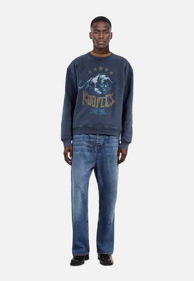 Marineblauwe sweatshirt met grafisch pantherontwerp en gouden sterren, gecombineerd met rechte spijkerbroek in blauw en zwarte schoenen.