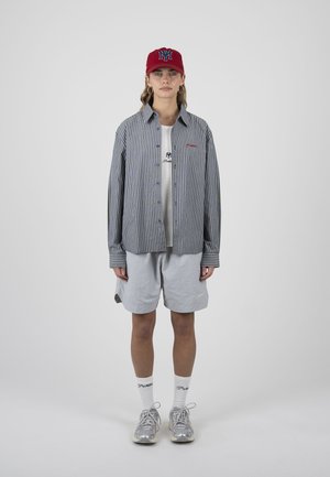 Frau steht nach vorne gerichtet, trägt eine rote Kappe, ein gestreiftes Hemd mit Knöpfen über einem weißen T-Shirt, graue Shorts, weiße Socken und graue Sneakers vor einfachem Hintergrund.