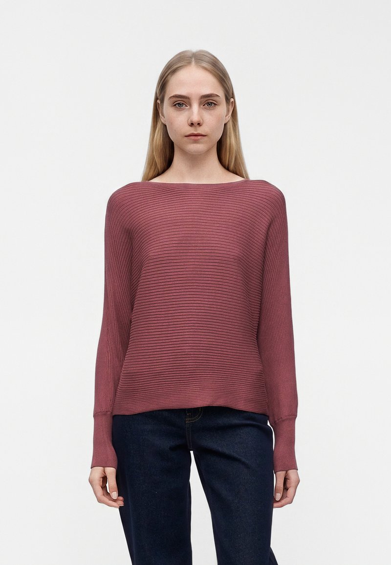 Jeune femme aux longs cheveux blonds portant un pull mauve côtelé à manches longues et un jean bleu foncé, debout devant un fond blanc uni.