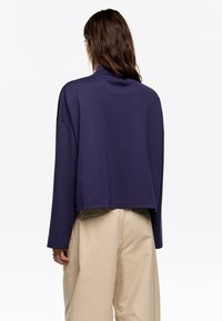 Paarse cropped sweatshirt met een hoge hals en lange mouwen, gecombineerd met losse beige broek, weergegeven tegen een eenvoudige witte achtergrond.