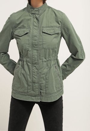 Femme portant une veste militaire verte zippée avec poches à rabat sur la poitrine et poches latérales zippées, associée à un pantalon noir.