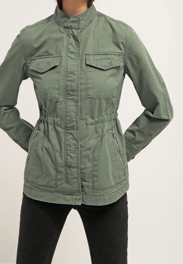 Femme portant une veste militaire verte zippée avec poches à rabat sur la poitrine et poches latérales zippées, associée à un pantalon noir.