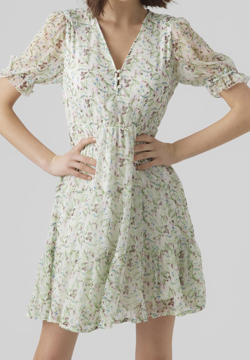 Robe fleurie en tissu vert clair avec des manches courtes bouffantes, un décolleté en V et un ourlet à volants. Présente un motif délicat de fleurs roses et bleues.