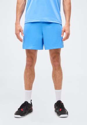 Mężczyzna ubraný w jasnoniebieskie sportowe szorty z siateczki, białe skarpetki i czarne buty sportowe z czerwonymi akcentami, stoi z rękami swobodnie opuszczonymi po bokach.