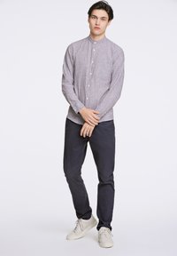 Camicia grigia chiaro con colletto stretto, maniche lunghe e taschino sul petto, abbinata a pantaloni blu scuro e scarpe da ginnastica chiare.