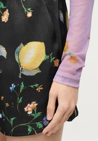 Χέρι που ακουμπά κοντά στο μηρό, φοράει μαύρη φούστα με κίτρινες λεμονιές και λουλουδάτα σχέδια, σε συνδυασμό με ένα μωβ τοπ μακρυμάνικο με μικρά πορτοκαλί λουλούδια.