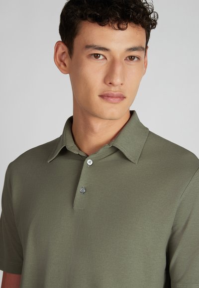 Jeune homme aux cheveux courts et bouclés portant un polo vert olive, se tenant devant un fond clair, légèrement tourné vers la droite.