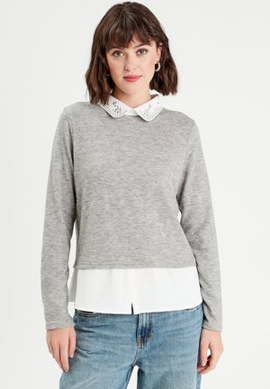 Graues Langarmoberteil mit einem weißen, gestickten Kragen, das über einem weißen Hemdsaum getragen wird, kombiniert mit hellblauen, locker sitzenden Jeans.