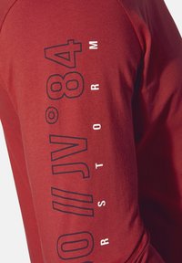Red long-sleeve shirt featuring large printed text in dark navy. Soft cotton material with a fitted design and round neckline.