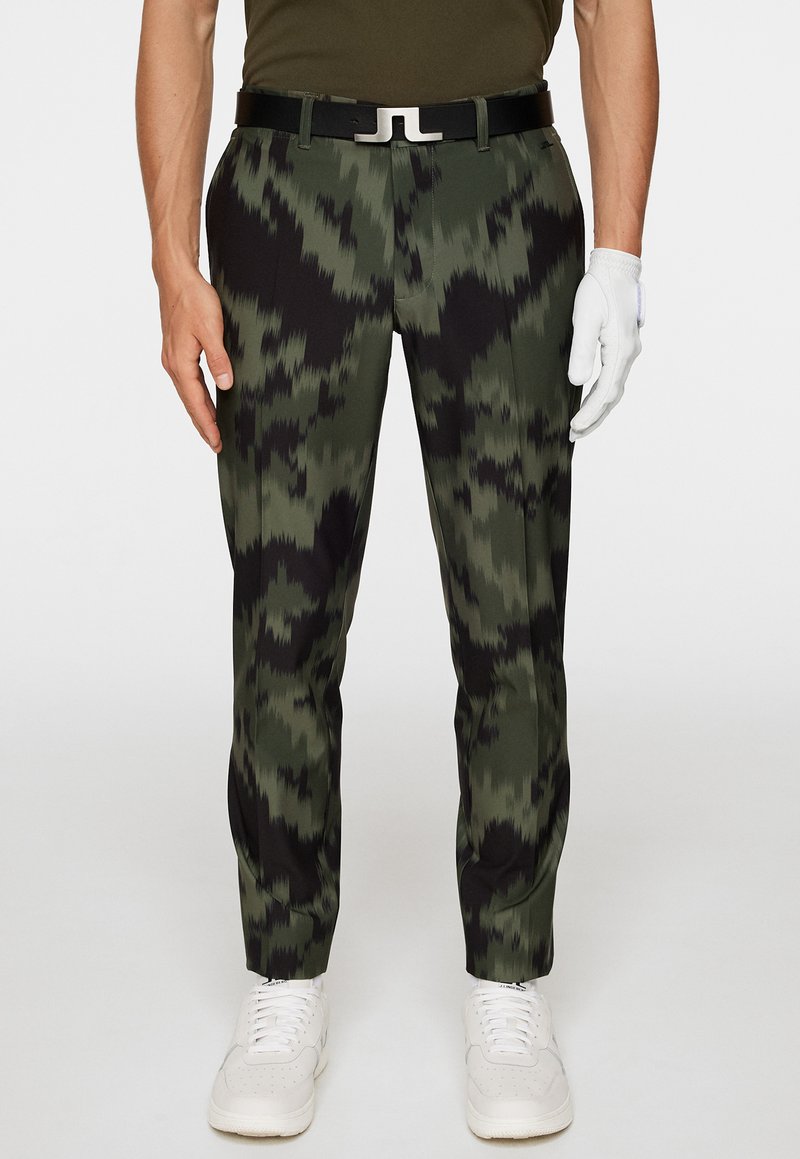 Kamouflage-mönstrade byxor i grön och svart; gjorda av ett glatt tyg; kombinerade med ett svart bälte med en silverspänne; vita skor.