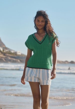 Ung kvinde med krøllet hår iført en grøn strikket top og stribede shorts går på en strand med bølger og bakker i baggrunden.