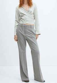 Top avvolgente verde chiaro con maniche lunghe e dettaglio di annodatura frontale, abbinato a pantaloni grigi a gamba larga con tasche frontali e una texture liscia.